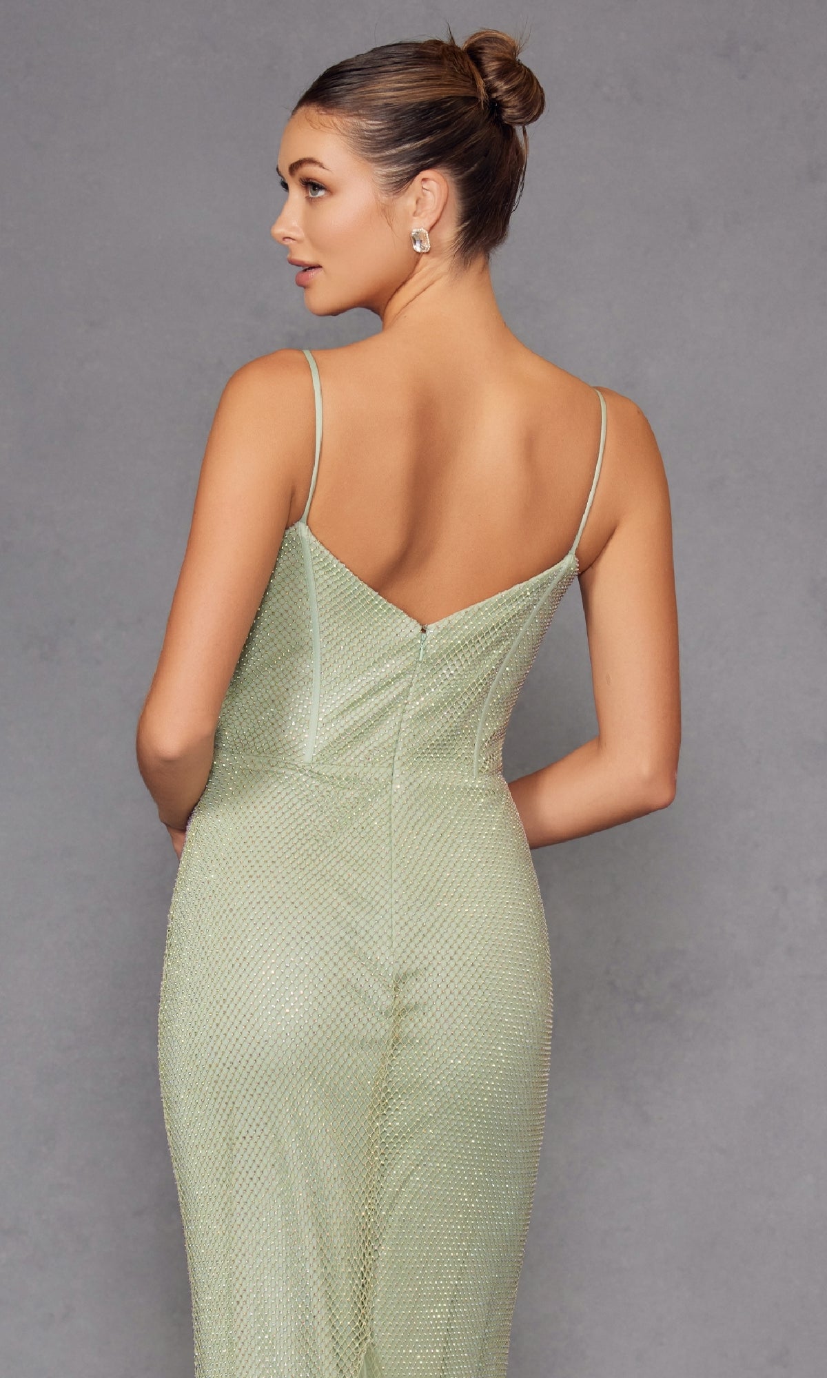 A woman in a pale green, shimmery Long Corset Beaded Prom Dress: Juliet JT2470S with thin straps stands with her back to the camera against a gray background, highlighting the dress’s elegant and beaded formal design.